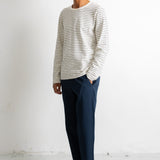 A man wearing a cream and navy striped long-sleeve knit sweater, navy blue trousers, and black and white canvas sneakers, standing in profile against a white studio wall.