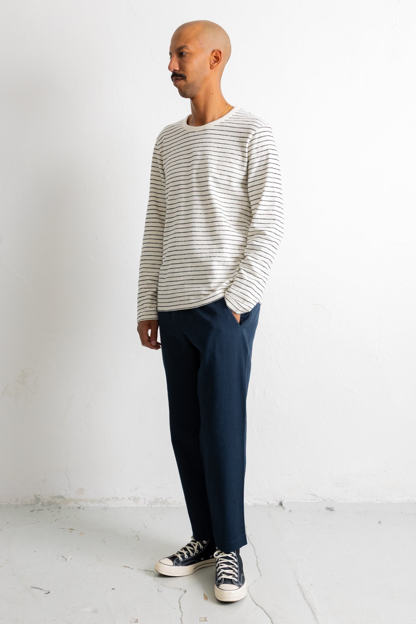 A man wearing a cream and navy striped long-sleeve knit sweater, navy blue trousers, and black and white canvas sneakers, standing in profile against a white studio wall.