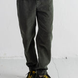 A person shown from the waist down wearing dark olive green corduroy pants, a white shirt, a navy jacket, and olive green sneakers with bright yellow laces, standing against a white wall.