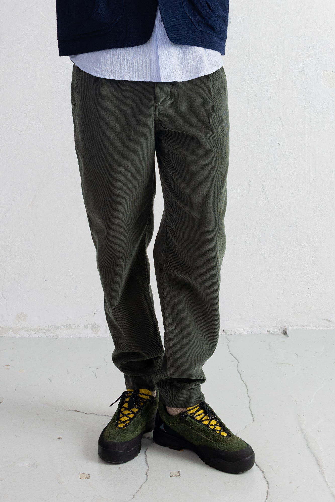 A person shown from the waist down wearing dark olive green corduroy pants, a white shirt, a navy jacket, and olive green sneakers with bright yellow laces, standing against a white wall.