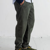 A person wearing olive green corduroy trousers, a navy blazer, a white shirt, and olive green lace-up shoes with yellow laces, standing against a white wall.
