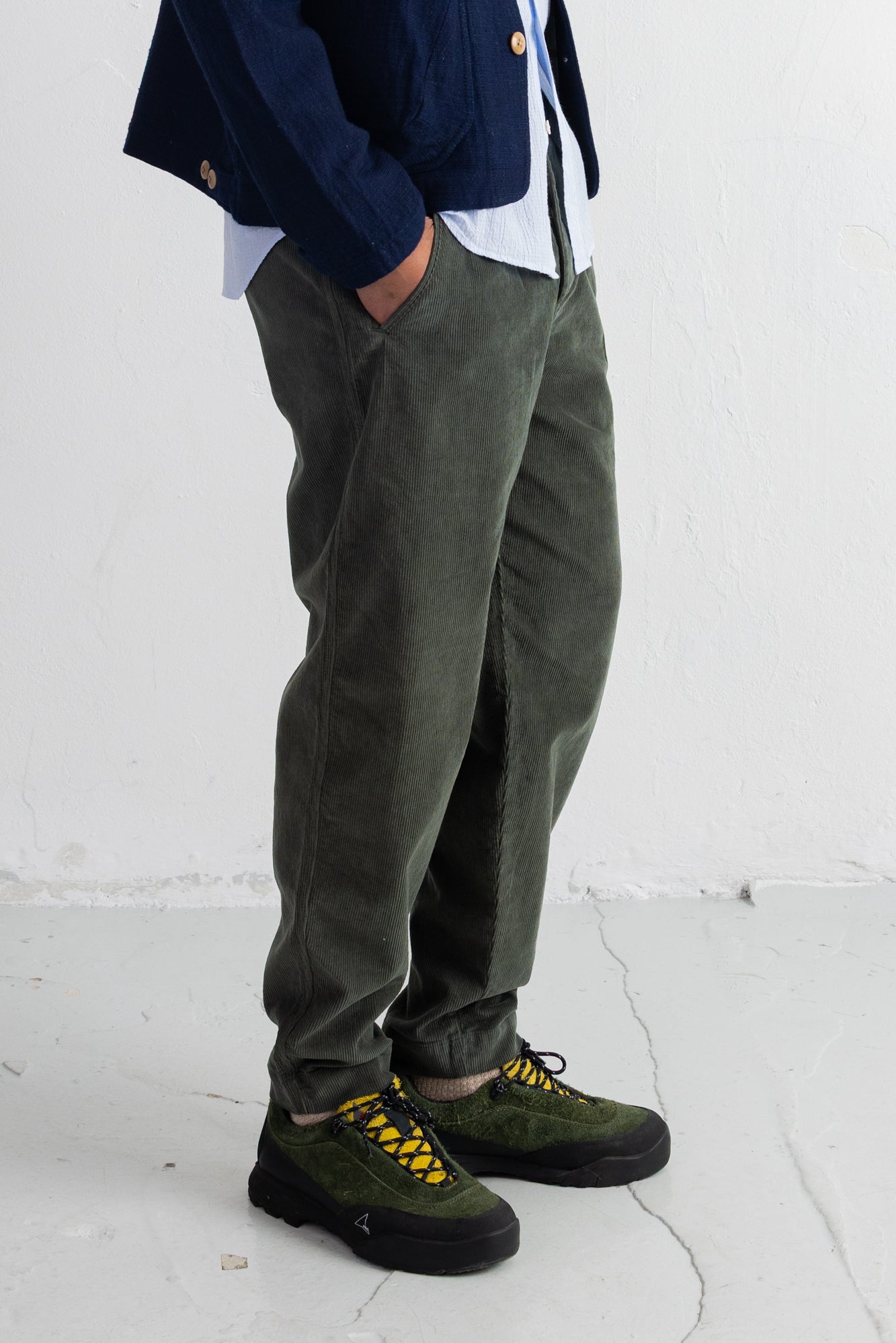 A person wearing olive green corduroy trousers, a navy blazer, a white shirt, and olive green lace-up shoes with yellow laces, standing against a white wall.