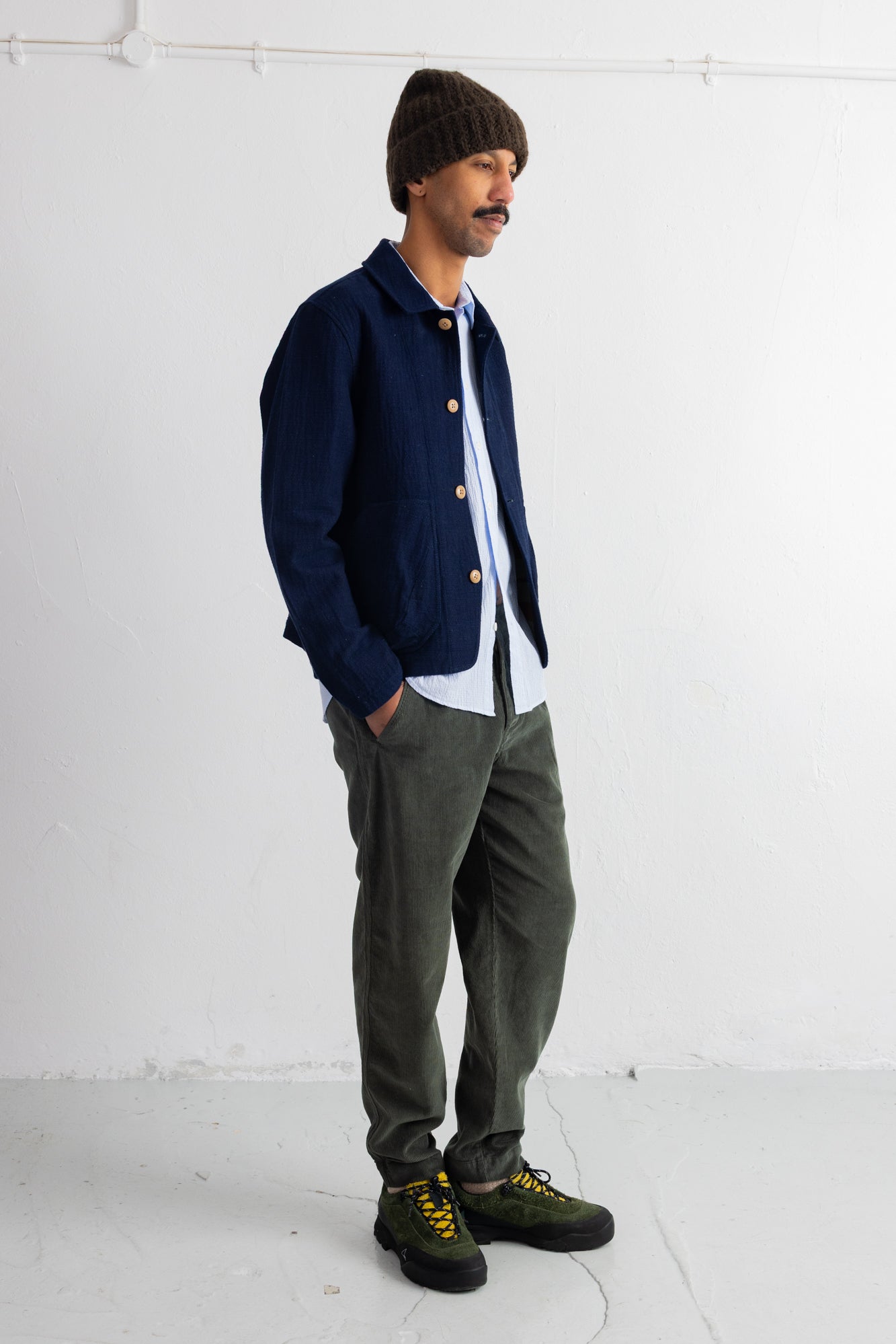 A man wearing a navy blue button-up cardigan over a white shirt, olive green pants, and black sneakers with yellow laces, standing against a white studio wall.