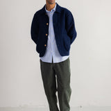 A man wearing a navy blue corduroy jacket over a light blue button-down shirt, paired with dark green trousers and black boots, standing against a plain white wall.