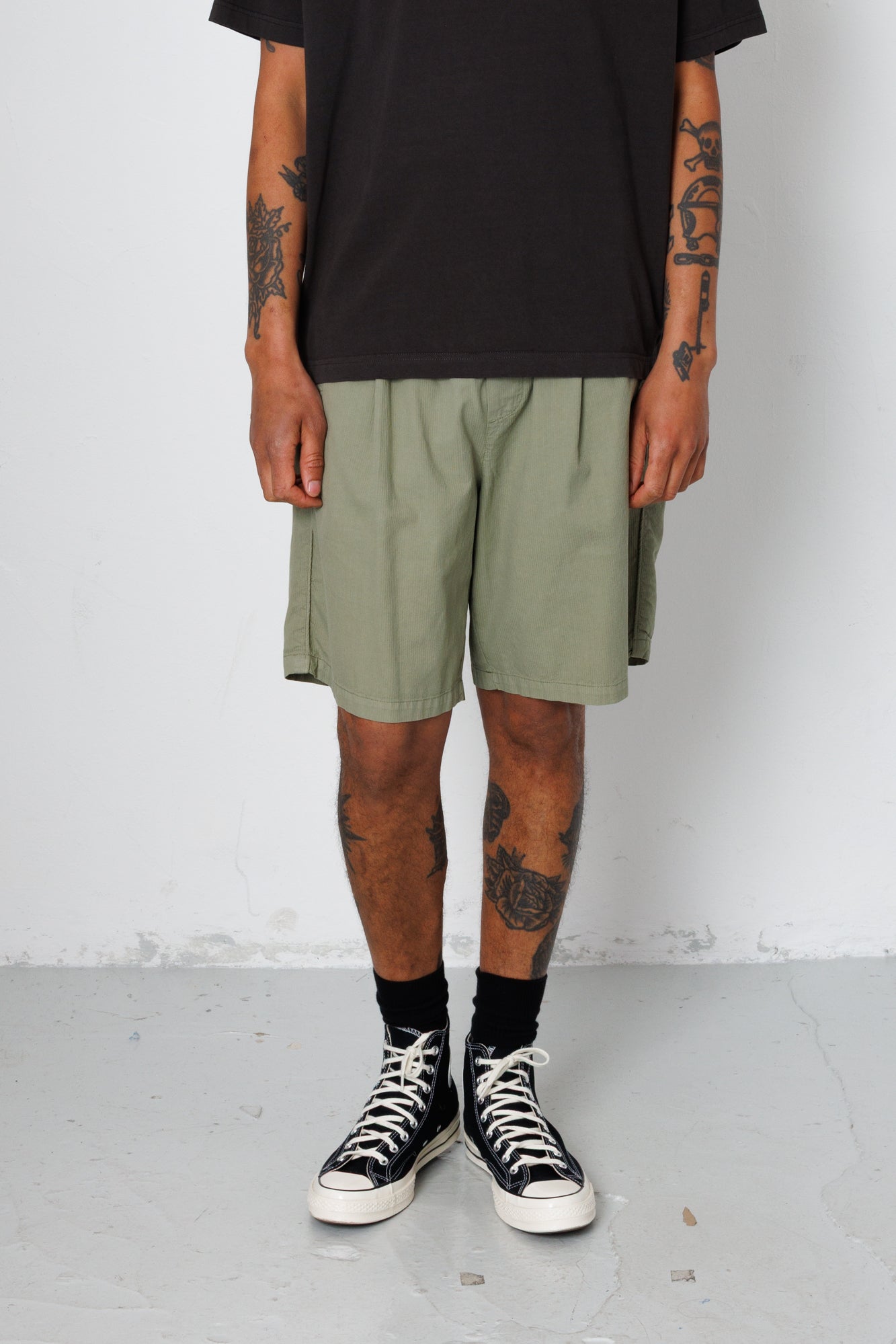 Person wearing a black t-shirt, green shorts, and black sneakers with white soles on a plain background.