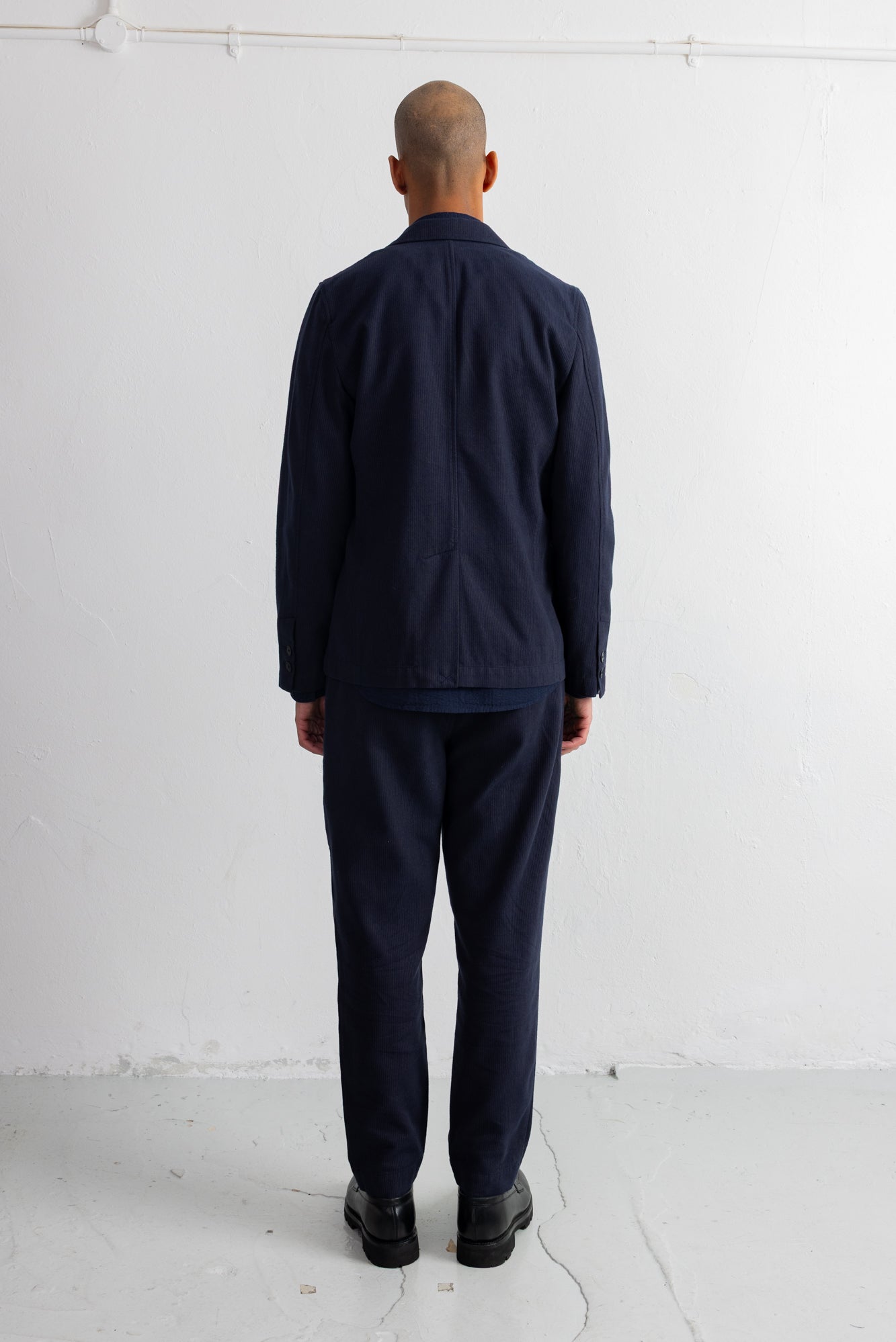 A man viewed from behind wearing a dark navy blue jacket and matching navy trousers, standing against a plain white wall.