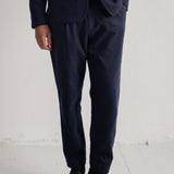 A person wearing dark navy blue trousers with a matching blazer and black leather shoes, standing against a plain off-white wall.