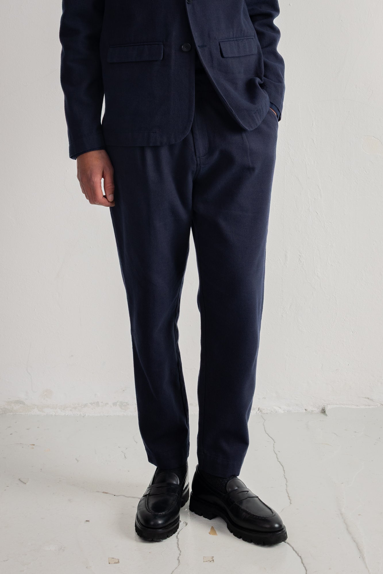A person wearing dark navy blue trousers with a matching blazer and black leather shoes, standing against a plain off-white wall.