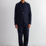 A man wearing a dark navy blue blazer and matching trousers over a dark shirt, standing against a plain white wall.