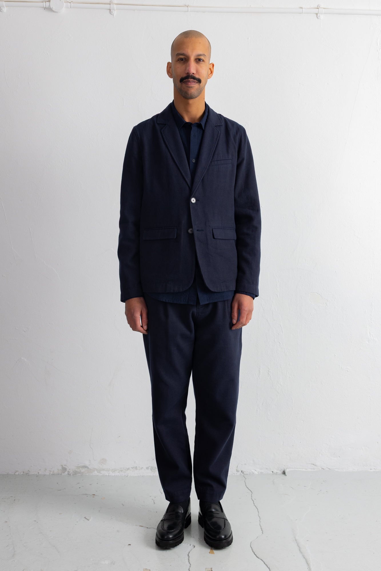 A man wearing a dark navy blue blazer and matching trousers over a dark shirt, standing against a plain white wall.