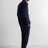 A man in a dark navy blue two-piece suit, consisting of a long-sleeve button-up shirt and matching trousers, standing in profile against a white studio wall.