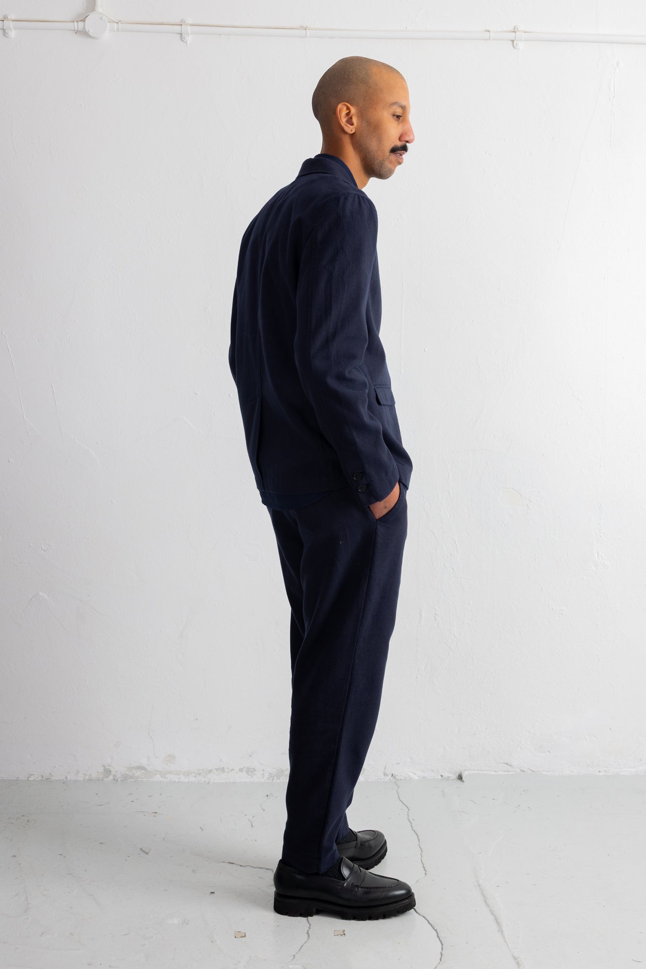 A man in a dark navy blue two-piece suit, consisting of a long-sleeve button-up shirt and matching trousers, standing in profile against a white studio wall.