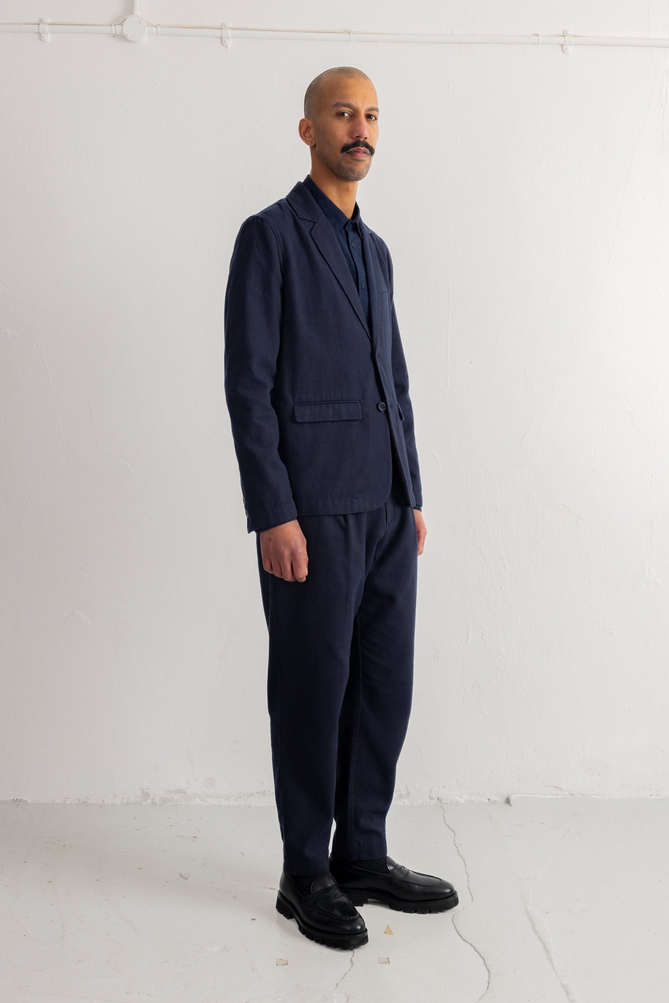 A man wearing a dark navy blue two-piece suit with a single-breasted blazer and matching trousers, paired with a dark shirt and black leather shoes, standing against a white studio background.