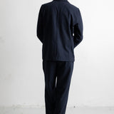 A man viewed from behind wearing a dark navy blue suit, consisting of a jacket and trousers, paired with dark brown shoes, standing against a white wall.