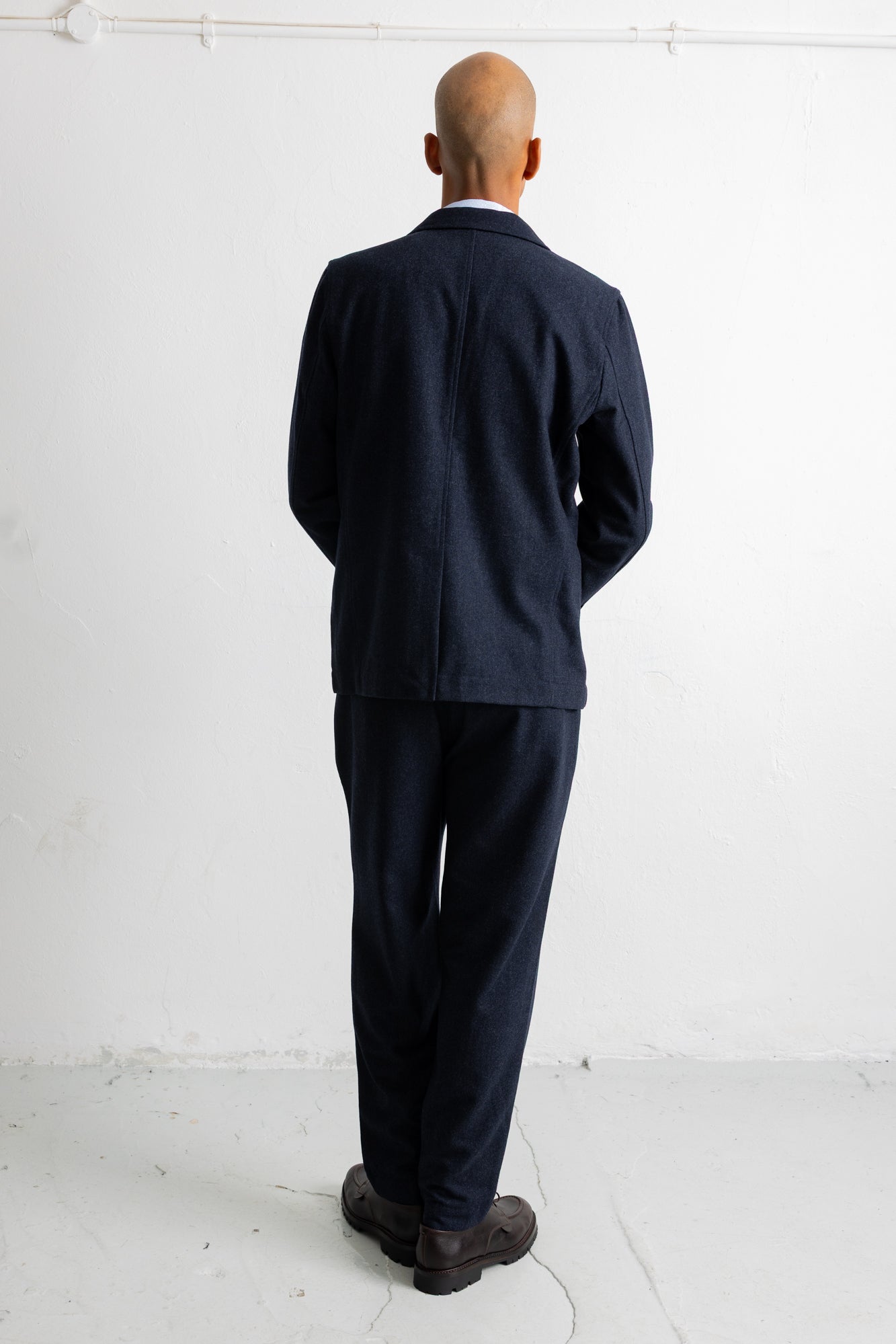 A man viewed from behind wearing a dark navy blue suit, consisting of a jacket and trousers, paired with dark brown shoes, standing against a white wall.