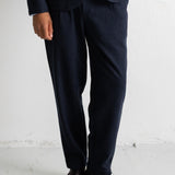 A person wearing dark navy trousers and a matching blazer, paired with dark brown leather shoes, standing against a white wall.