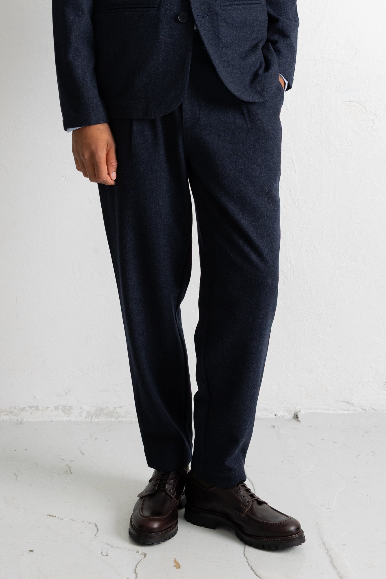 A person wearing dark navy trousers and a matching blazer, paired with dark brown leather shoes, standing against a white wall.