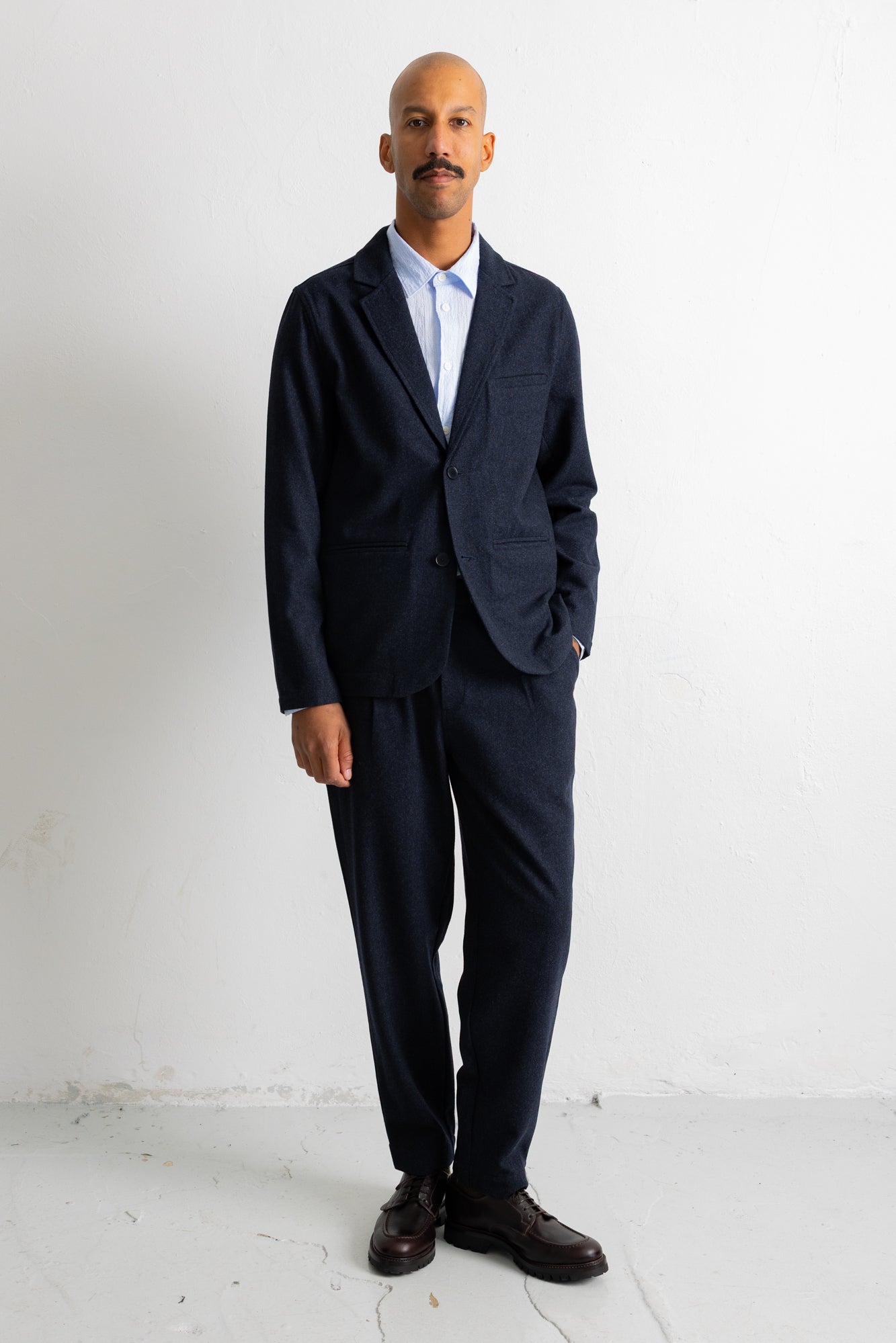 A man wearing a dark navy blue suit with a light blue dress shirt and dark brown leather shoes, standing against a white studio background.