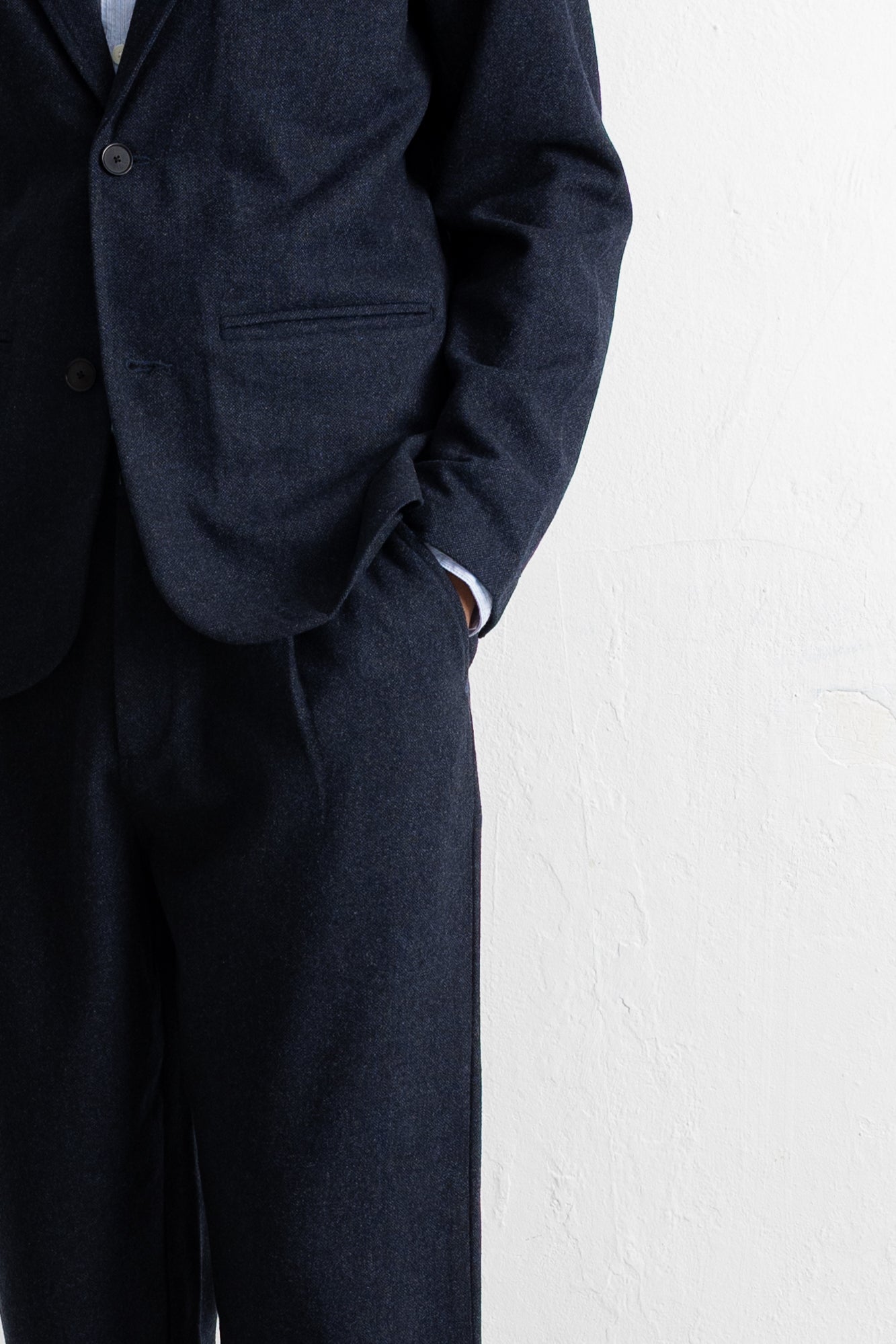 Close-up of a person wearing a dark navy wool suit, showing the jacket lapel, button, pocket, and trousers against a white textured wall.