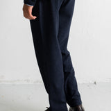 A person shown from the waist down wearing dark navy trousers and dark brown leather shoes, standing against a white wall.