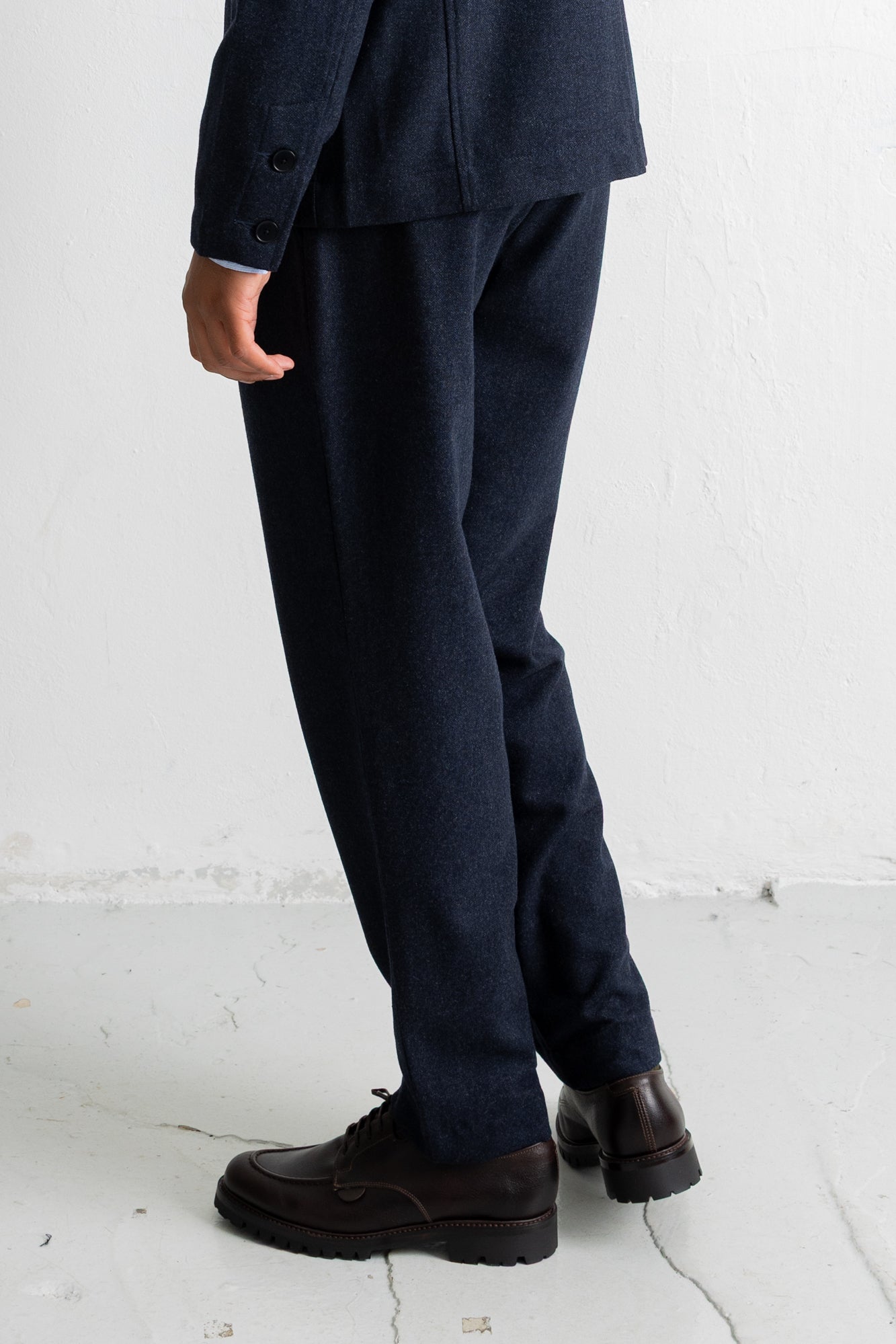 A person shown from the waist down wearing dark navy trousers and dark brown leather shoes, standing against a white wall.