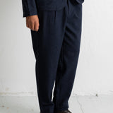 A person wearing dark navy pleated trousers with a matching blazer and dark brown leather shoes, standing against a white wall.