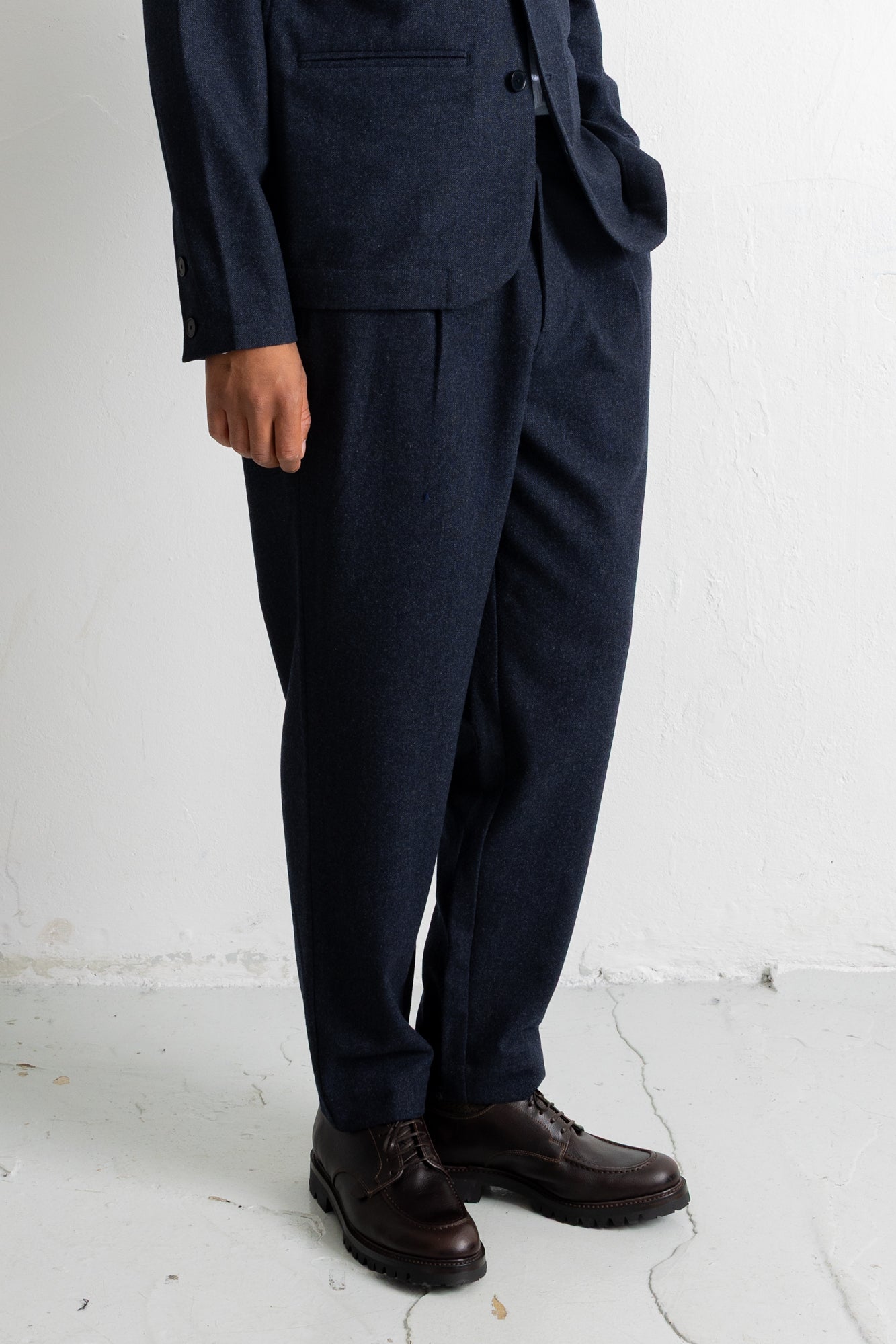 A person wearing dark navy pleated trousers with a matching blazer and dark brown leather shoes, standing against a white wall.
