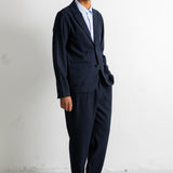 A man wearing a dark navy blue two-piece suit with a light blue shirt and dark brown dress shoes, standing against a white wall.