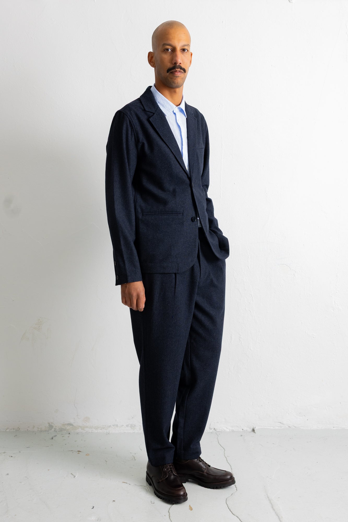 A man wearing a dark navy blue two-piece suit with a light blue shirt and dark brown dress shoes, standing against a white wall.