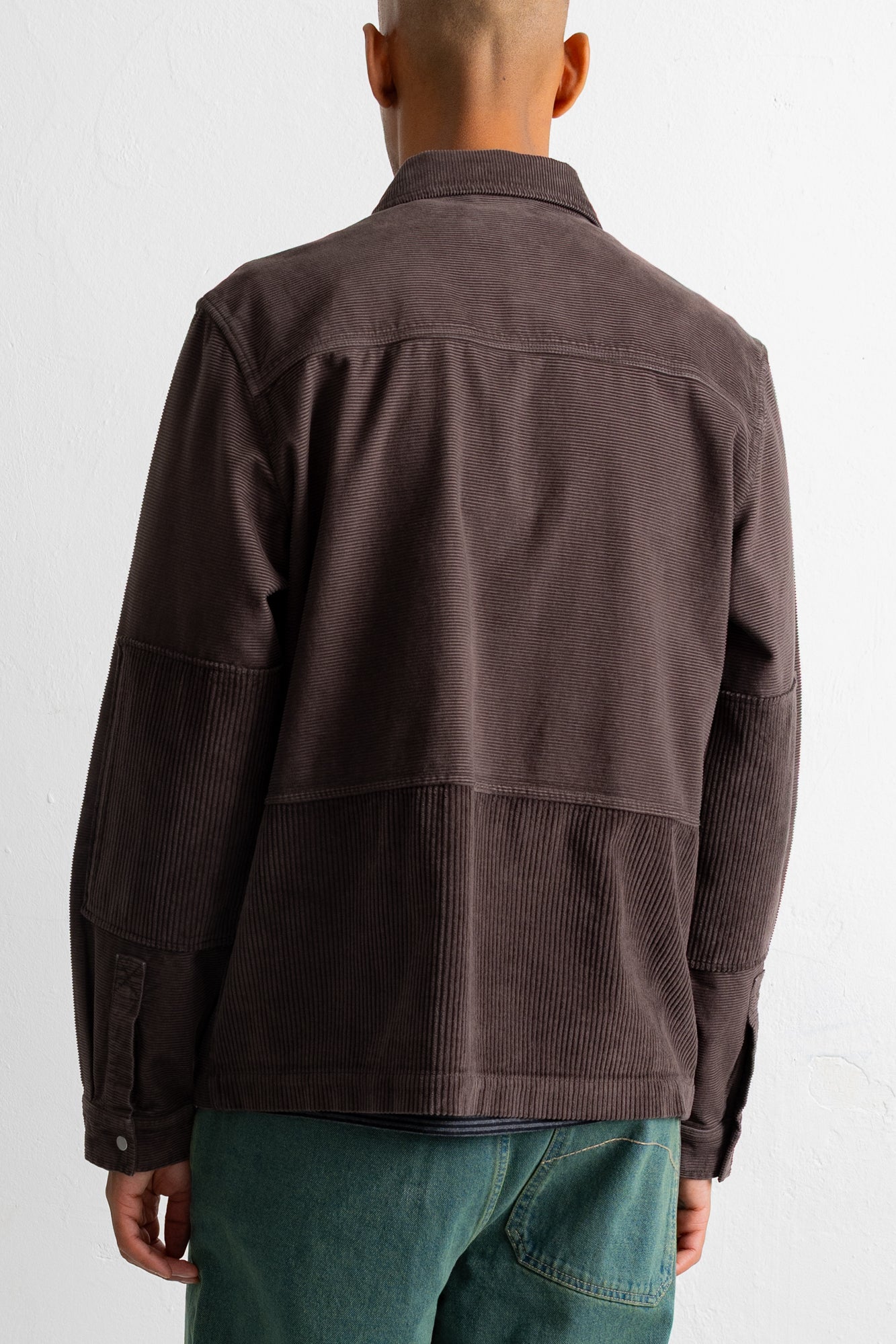 A man viewed from the back wearing a dark brown corduroy jacket and teal-green pants against a plain white background.