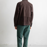 A man viewed from behind wearing a dark brown corduroy jacket, green trousers, and black-and-white sneakers, standing against a plain white studio wall.