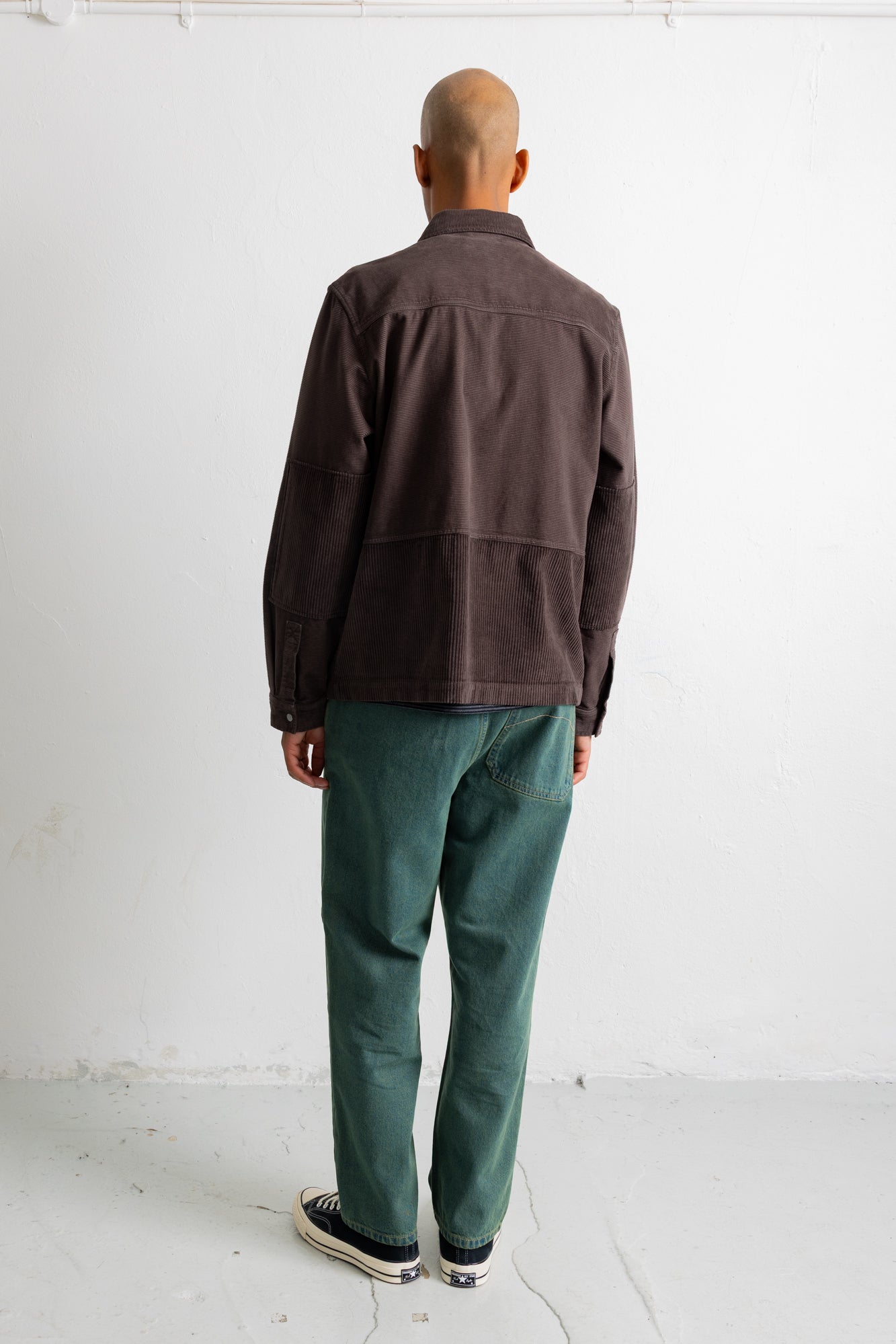 A man viewed from behind wearing a dark brown corduroy jacket, green trousers, and black-and-white sneakers, standing against a plain white studio wall.