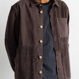 A man wearing a dark brown corduroy utility jacket over a navy blue striped t-shirt and teal pants, photographed against a plain white wall.