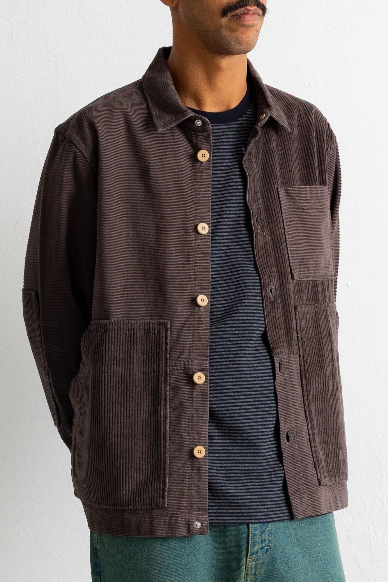 A man wearing a dark brown corduroy utility jacket over a navy blue striped t-shirt and teal pants, photographed against a plain white wall.