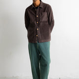 A man wearing a dark brown corduroy button-up shirt jacket, green trousers, and black-and-white sneakers, standing against a white wall.