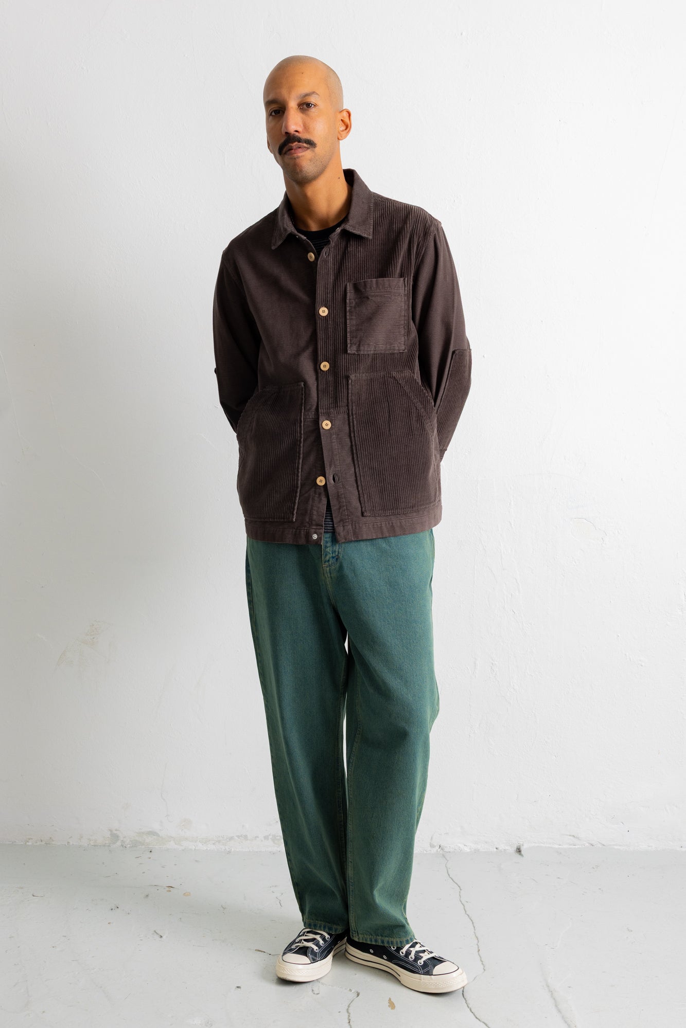 A man wearing a dark brown corduroy button-up shirt jacket, green trousers, and black-and-white sneakers, standing against a white wall.