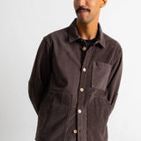 A man wearing a dark brown corduroy button-up shirt jacket over a black t-shirt and teal jeans, standing against a plain white wall.