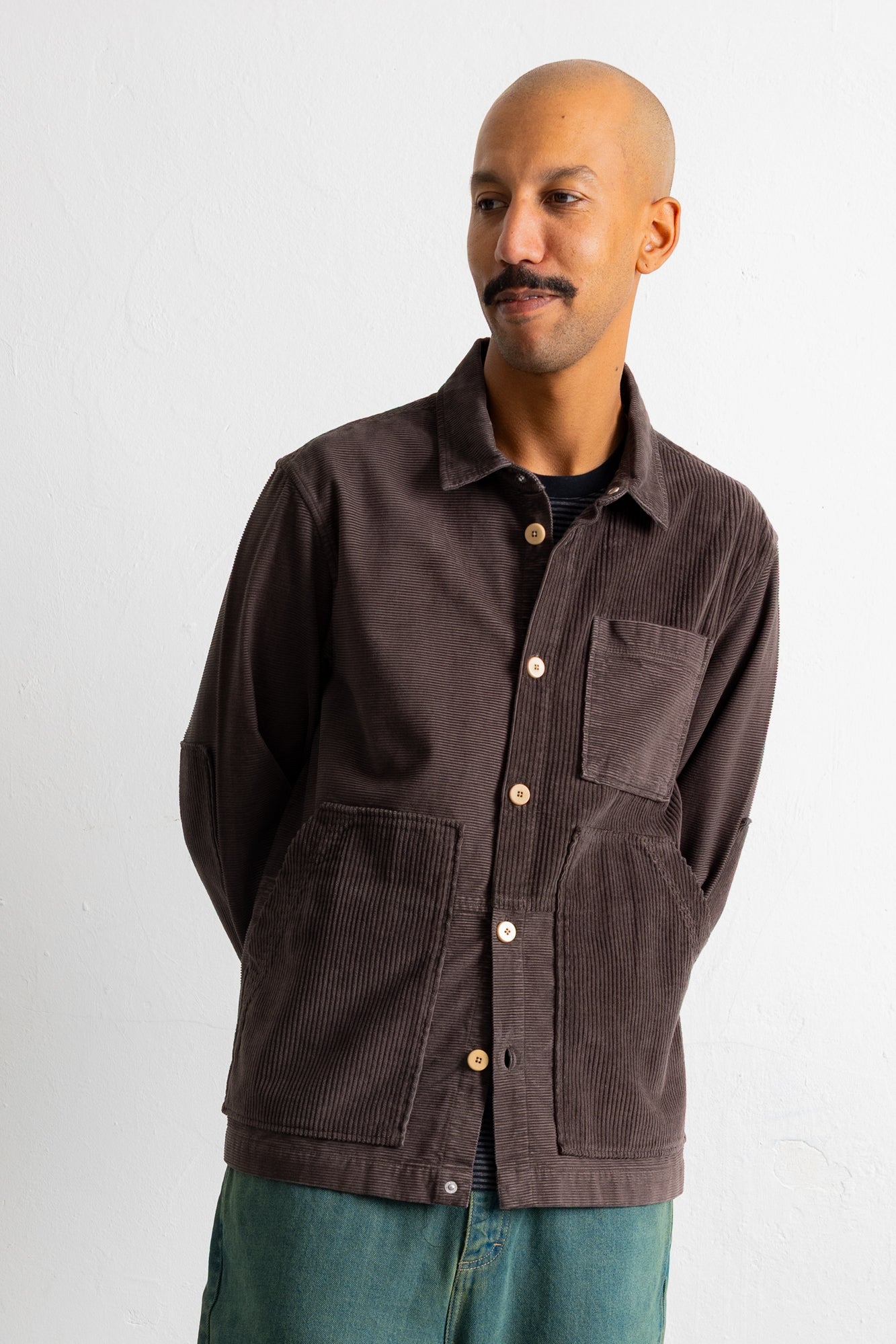 A man wearing a dark brown corduroy button-up shirt jacket over a black t-shirt and teal jeans, standing against a plain white wall.