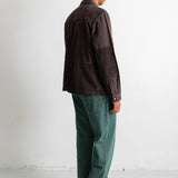 A man standing in profile wearing a dark brown patchwork jacket, green trousers, and black canvas sneakers against a plain white studio wall.