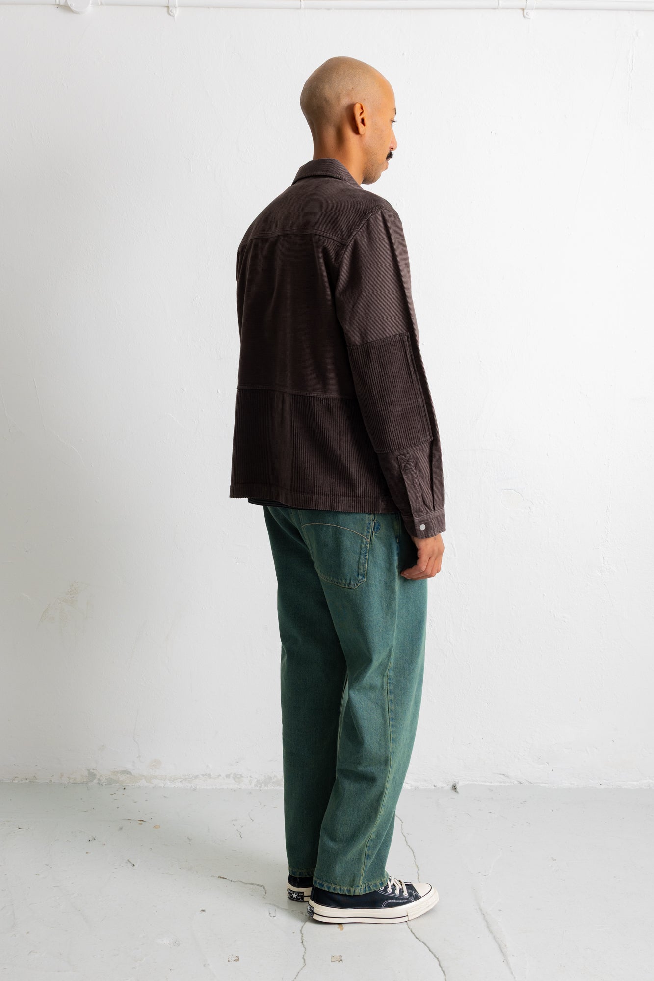A man standing in profile wearing a dark brown patchwork jacket, green trousers, and black canvas sneakers against a plain white studio wall.