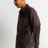 A man wearing a dark brown corduroy button-up shirt jacket and teal jeans, standing in profile against a plain off-white wall.