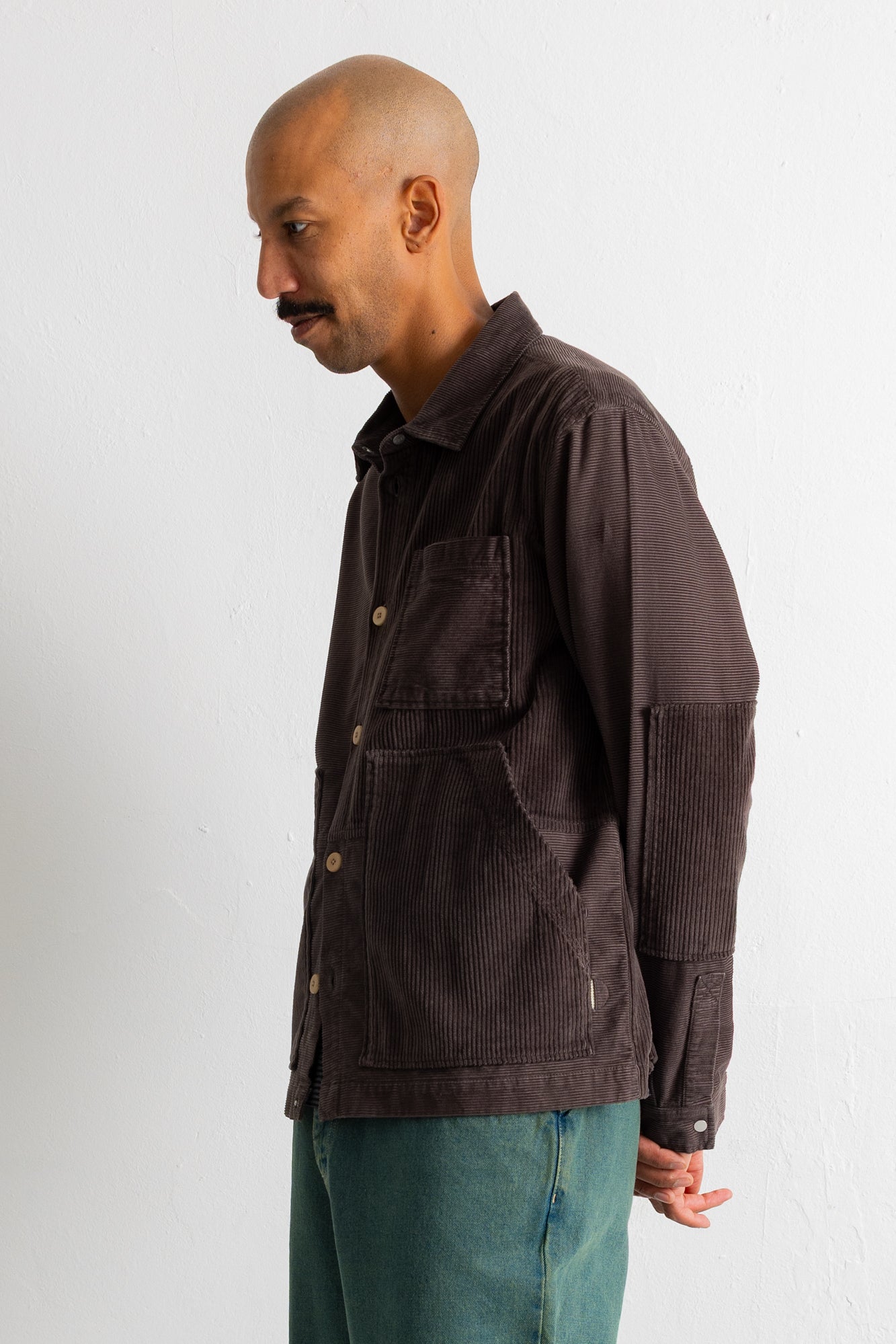 A man wearing a dark brown corduroy button-up shirt jacket and teal jeans, standing in profile against a plain off-white wall.