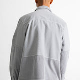 A man viewed from behind wearing a light gray patchwork jacket with corduroy details on the lower back and sleeves, paired with dark blue jeans against a plain white background.