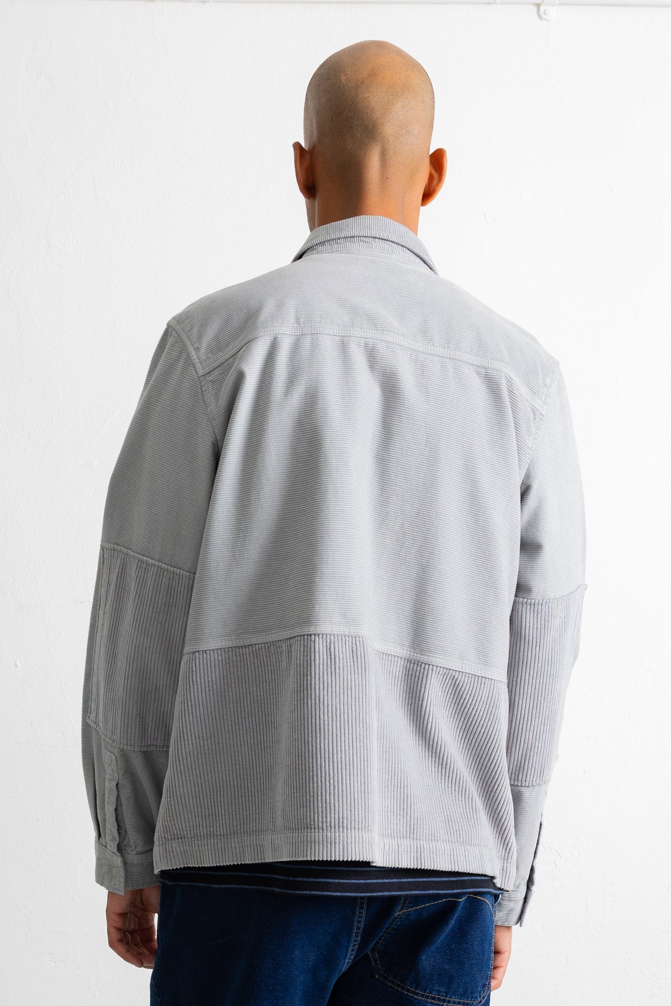 A man viewed from behind wearing a light gray patchwork jacket with corduroy details on the lower back and sleeves, paired with dark blue jeans against a plain white background.