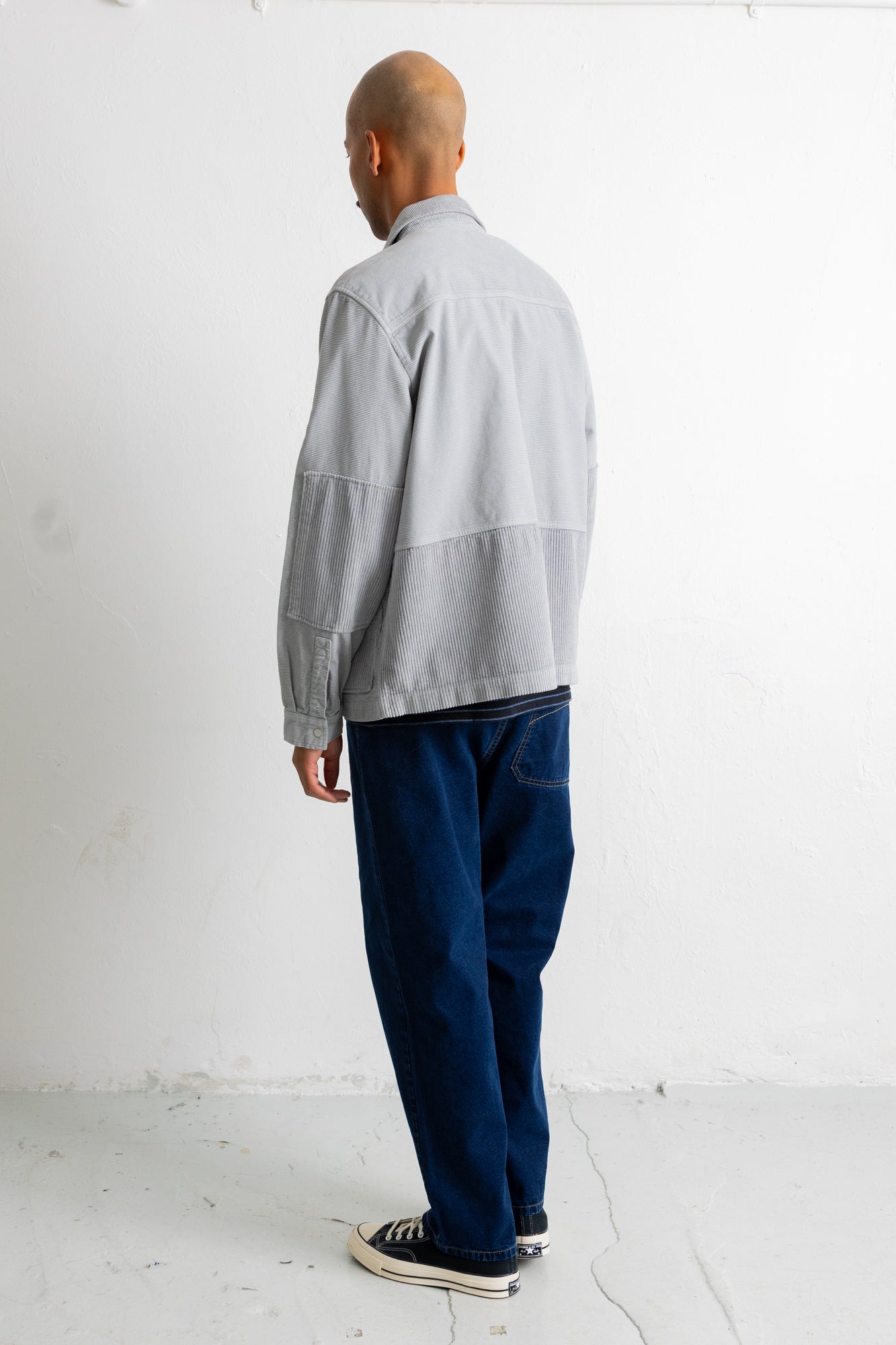 A man is shown from behind wearing a light gray long-sleeve overshirt, dark blue jeans, and black canvas sneakers, standing against a white studio wall.