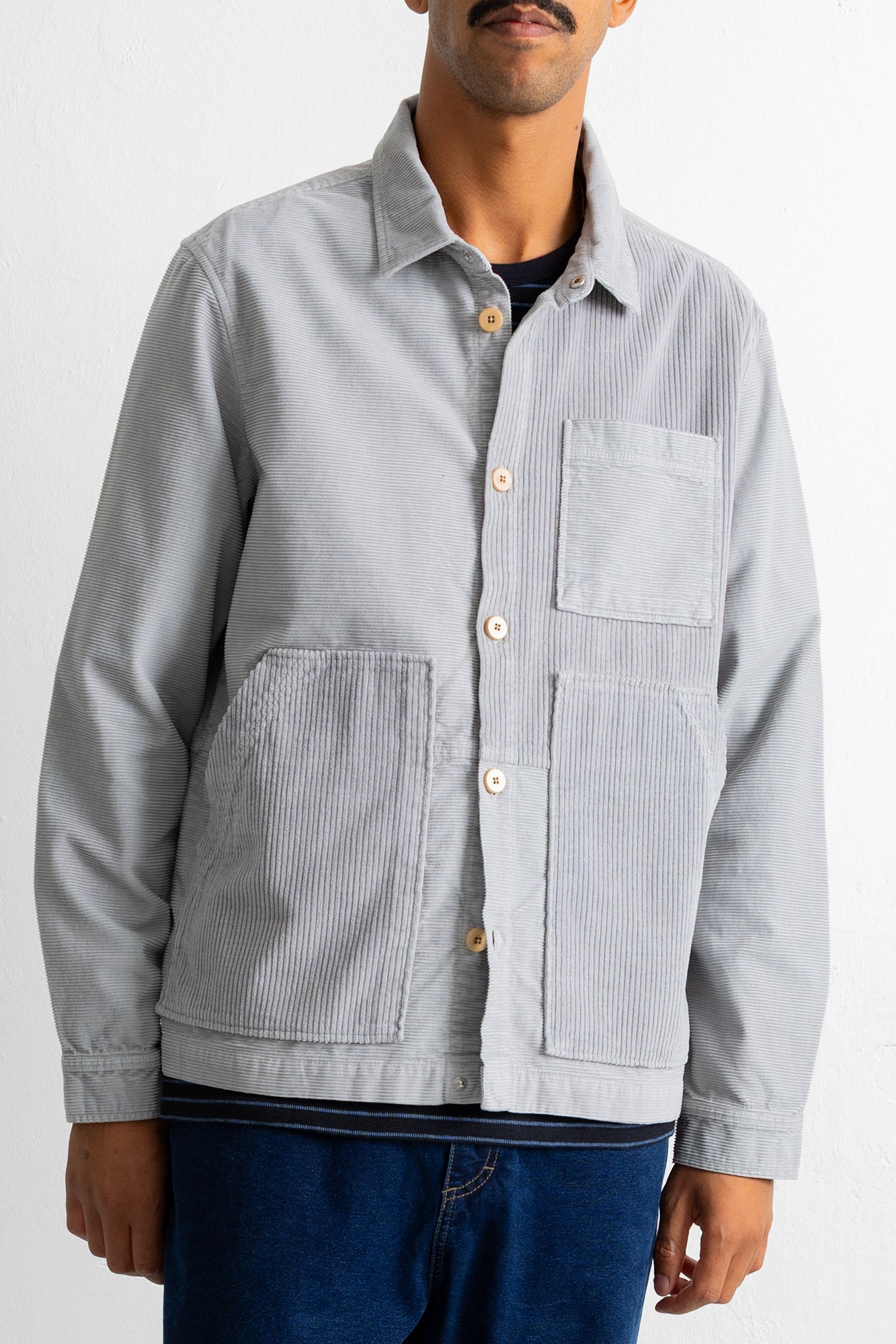A man wearing a light gray corduroy button-up overshirt with a chest pocket and two large front patch pockets, paired with dark blue jeans.