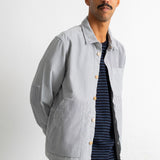 A man wearing a light gray corduroy utility jacket over a navy blue striped t-shirt and blue jeans, standing against a plain white studio background.