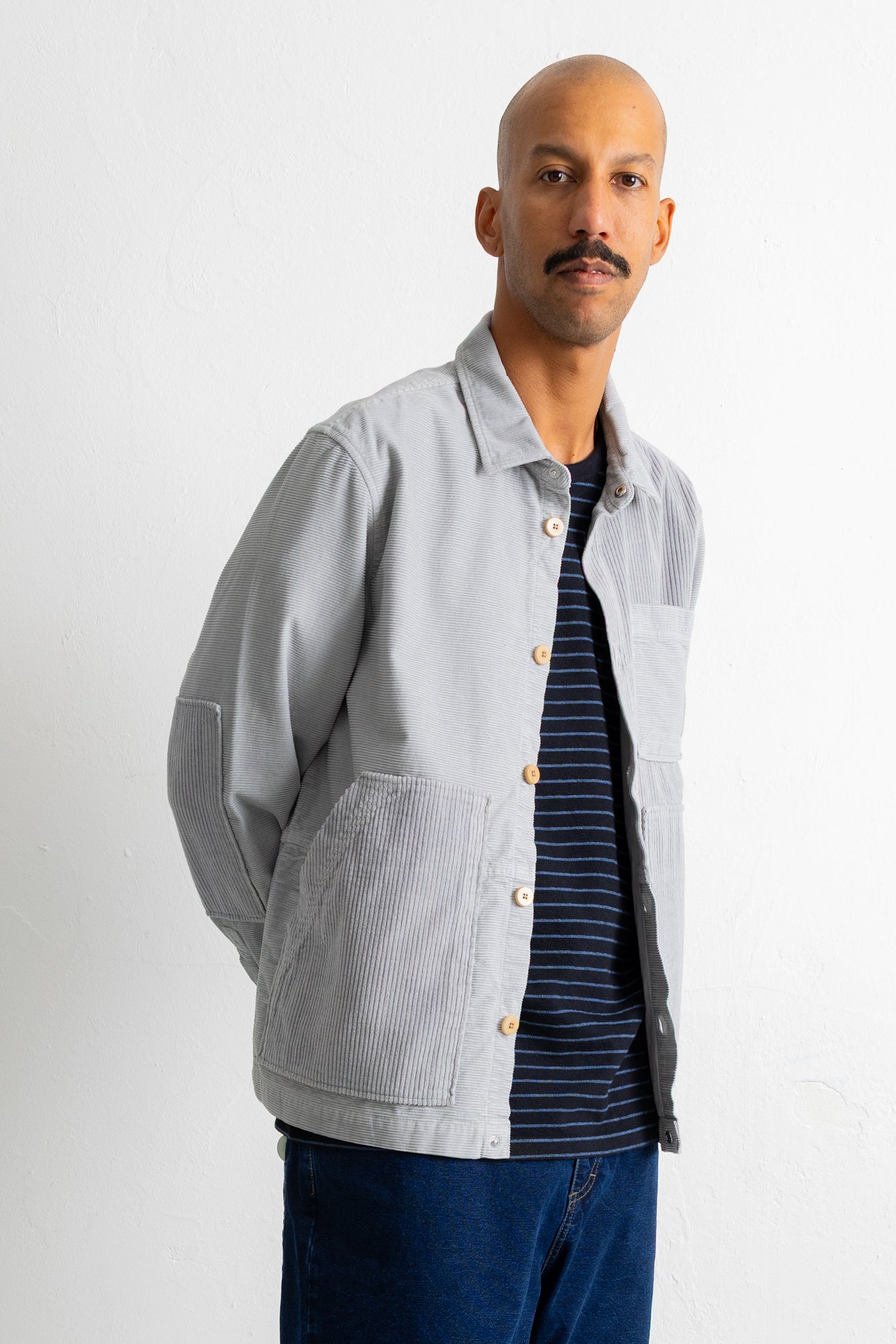 A man wearing a light gray corduroy utility jacket over a navy blue striped t-shirt and blue jeans, standing against a plain white studio background.