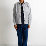 A man wearing a light gray button-up overshirt over a black and white striped shirt, dark blue jeans, and black and white sneakers, standing against a white wall.