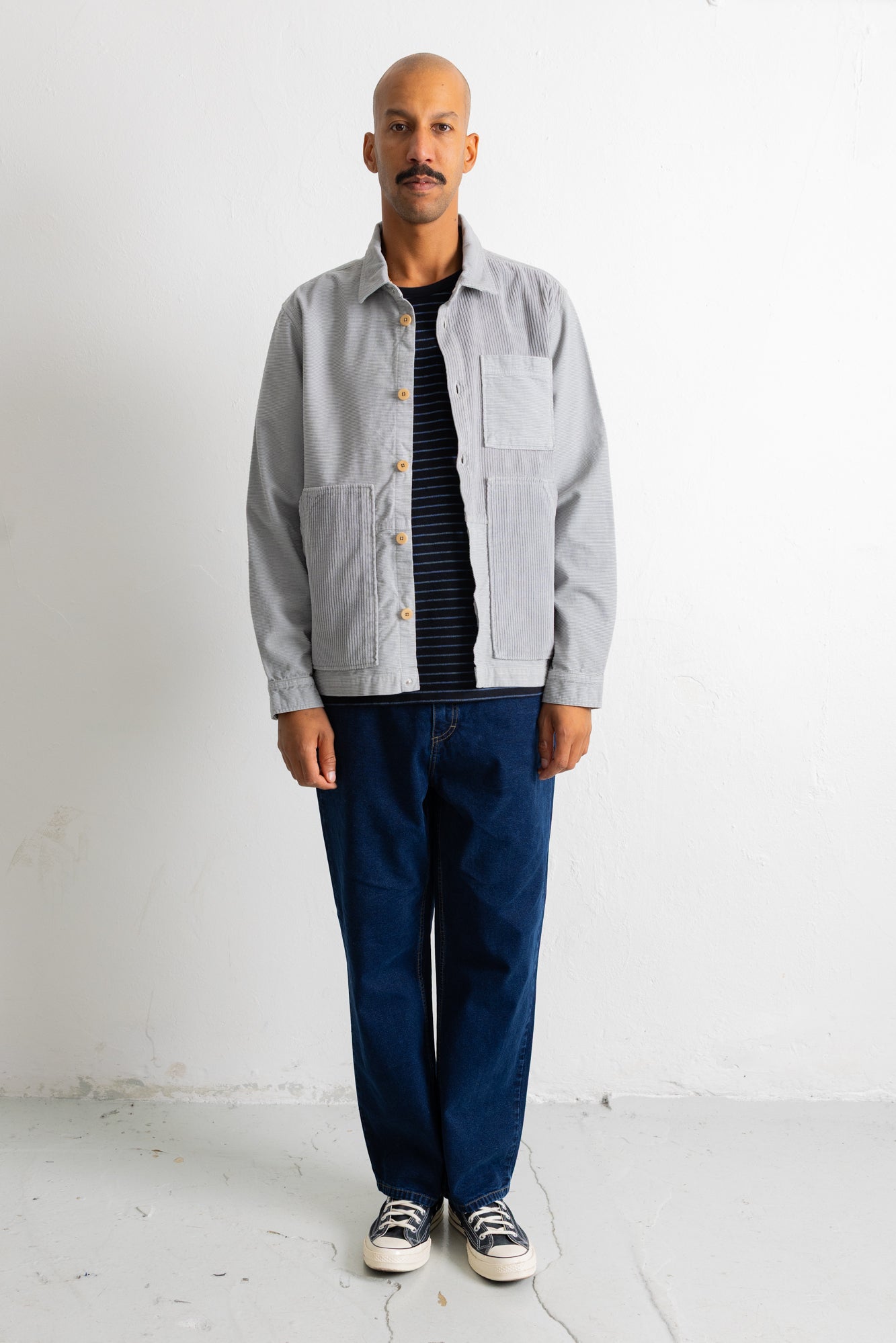 A man wearing a light gray button-up overshirt over a black and white striped shirt, dark blue jeans, and black and white sneakers, standing against a white wall.